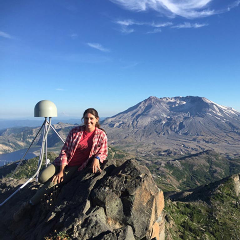 Maggie Holahan smiling at the camera from atop a rock formation with a mountain behind her