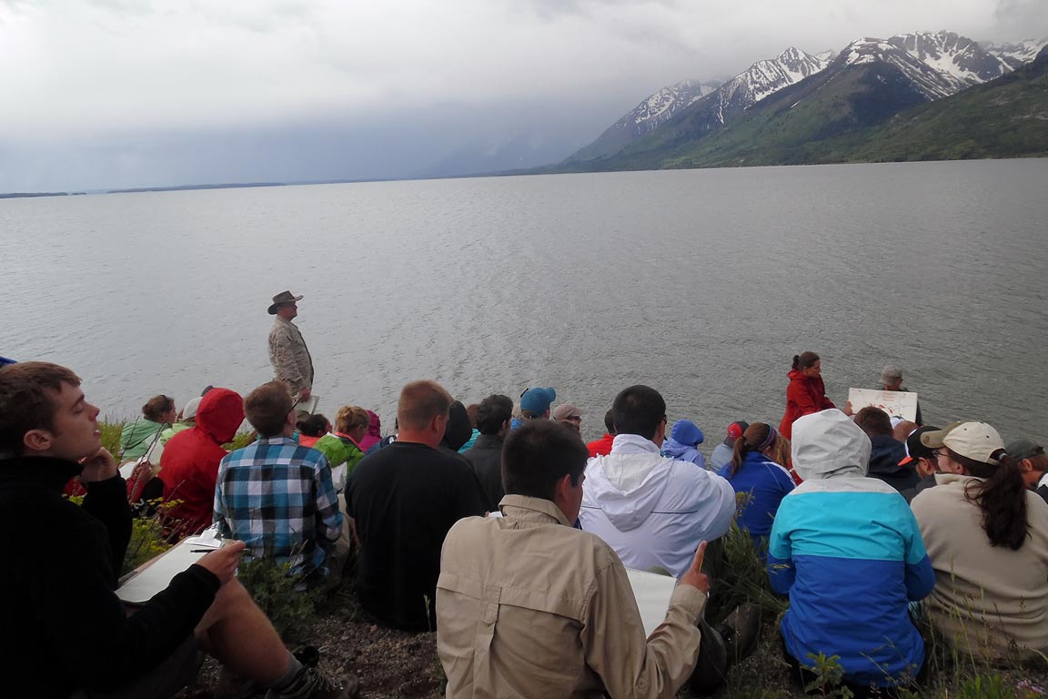 Field student sin Montana taking notes at the edge of a lake facing a mountain range
